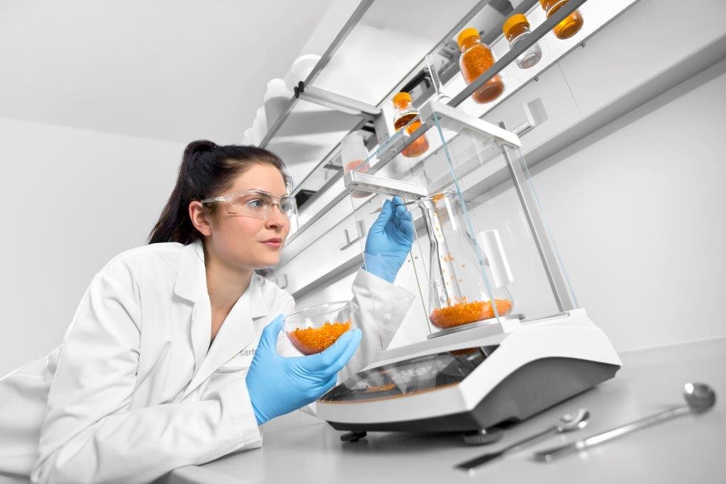 tecnica di laboratorio pesa granuli arancioni su bilancia con paravento